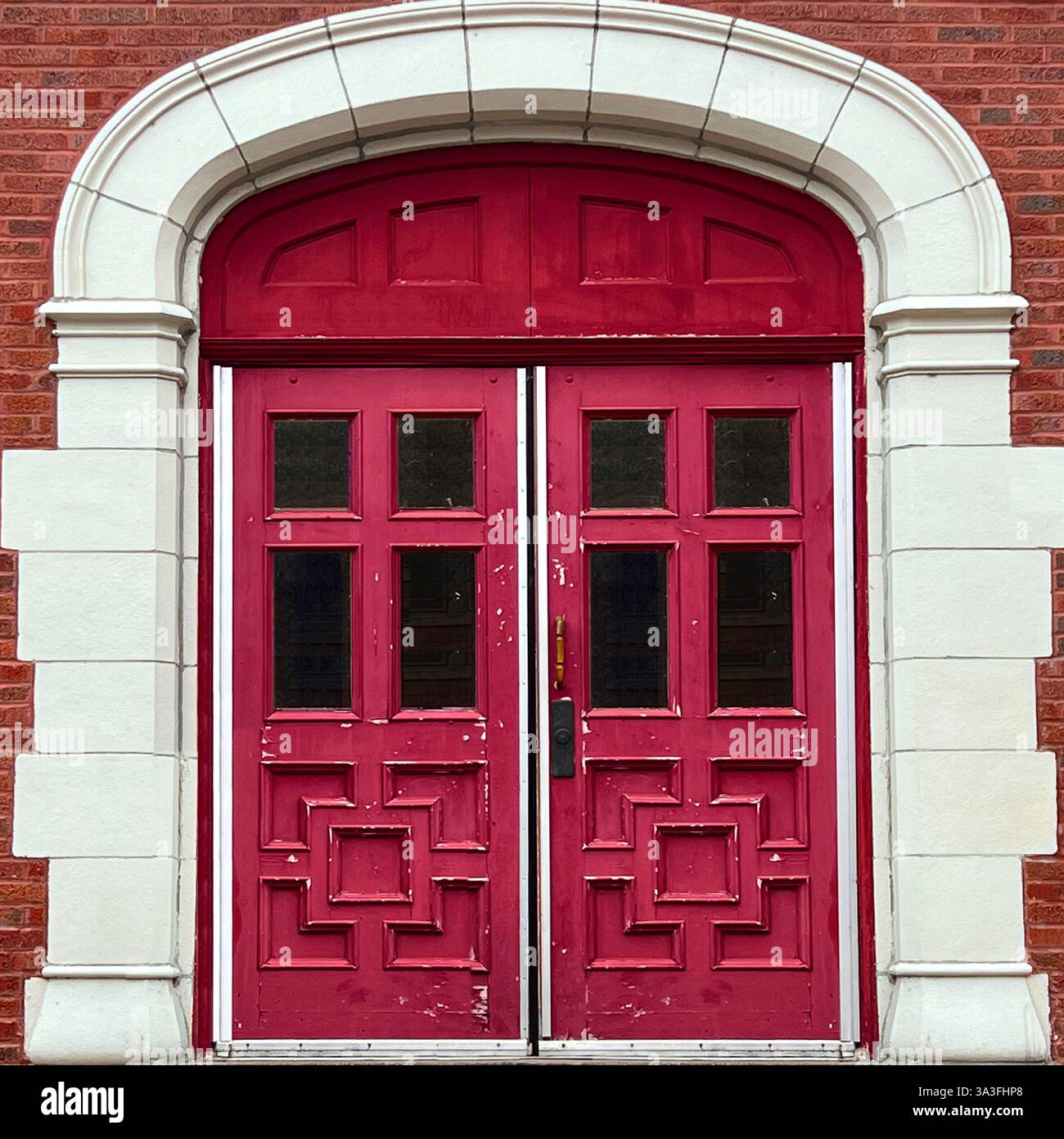 Weathered red double doors with intricate geometric panel designs, framed by a white stone archway against a red brick facade in Chicago. - Smartphone Captured Stock Image