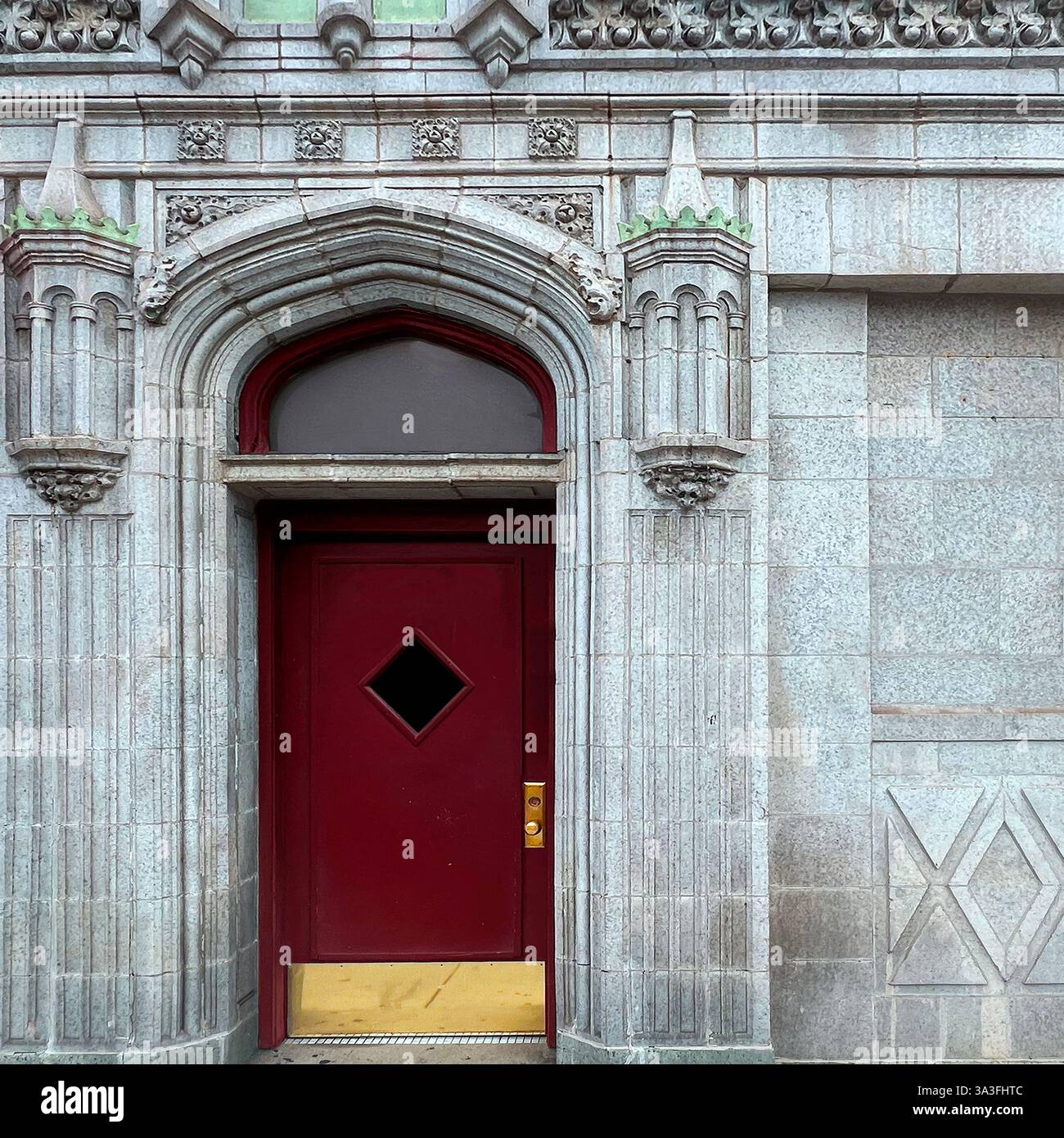 A Gothic-inspired red door with a diamond-shaped window, set within an ornate stone archway of a historic building. - Smartphone Captured Stock Image