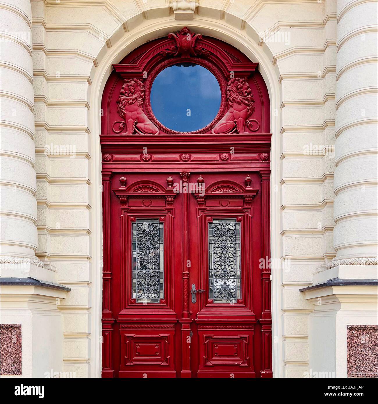 An ornate red double door with intricate ironwork, lion reliefs, and a circular transom, set within a grand historic building in Łódź, Poland - Smartphone Captured Stock Image