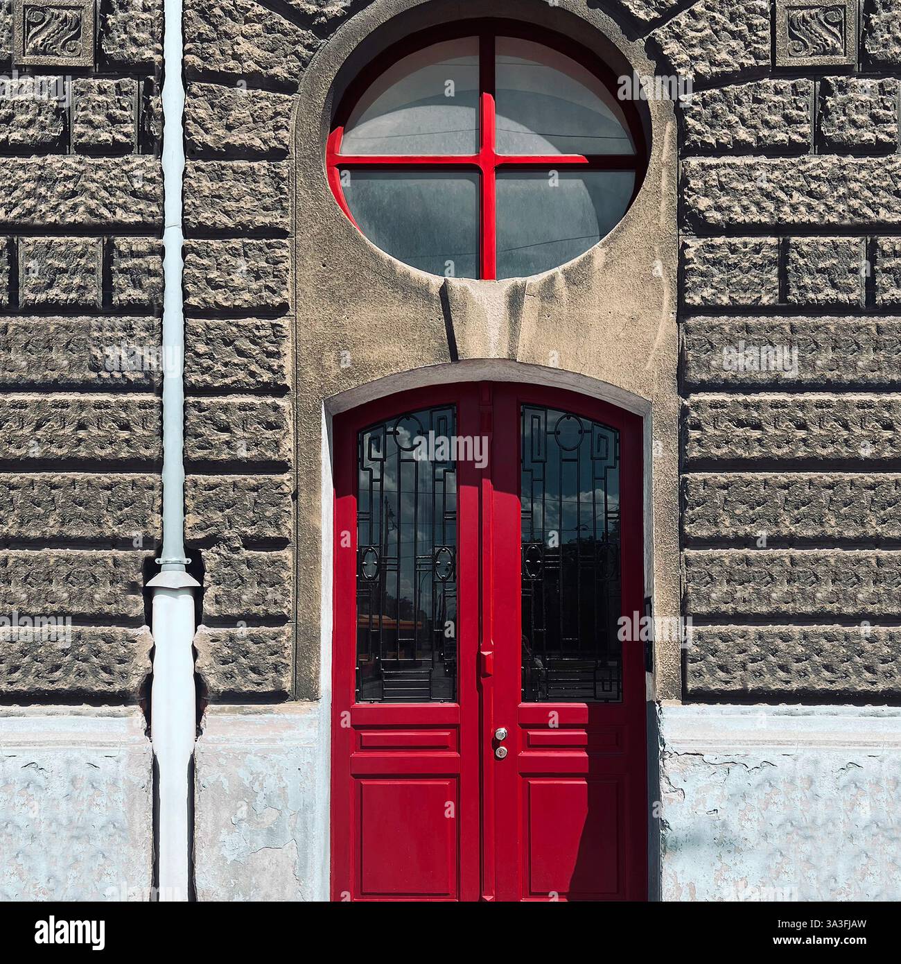 Red double door with intricate ironwork set in a textured stone facade, featuring a unique circular window above, in Łódź, Poland - Smartphone Captured Stock Image