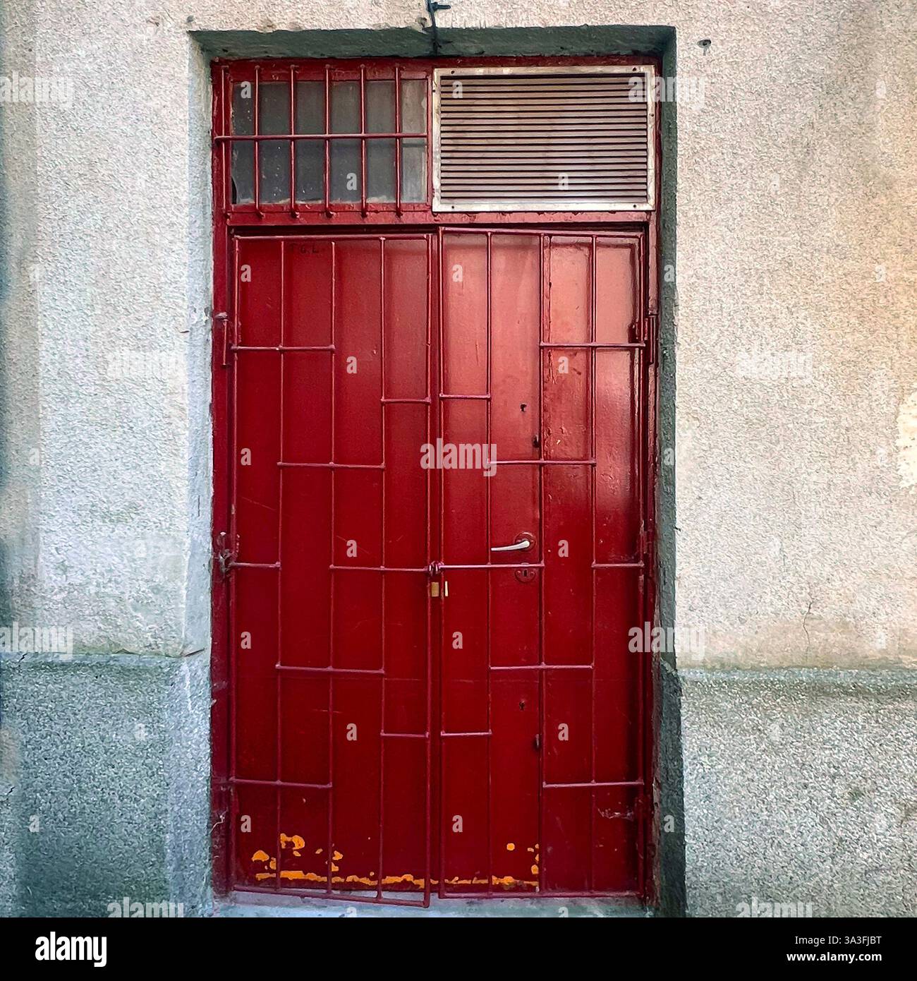 Red metal door with a security grid and ventilation panel, set in a textured concrete wall in Warsaw, Poland. - Smartphone Captured Stock Image