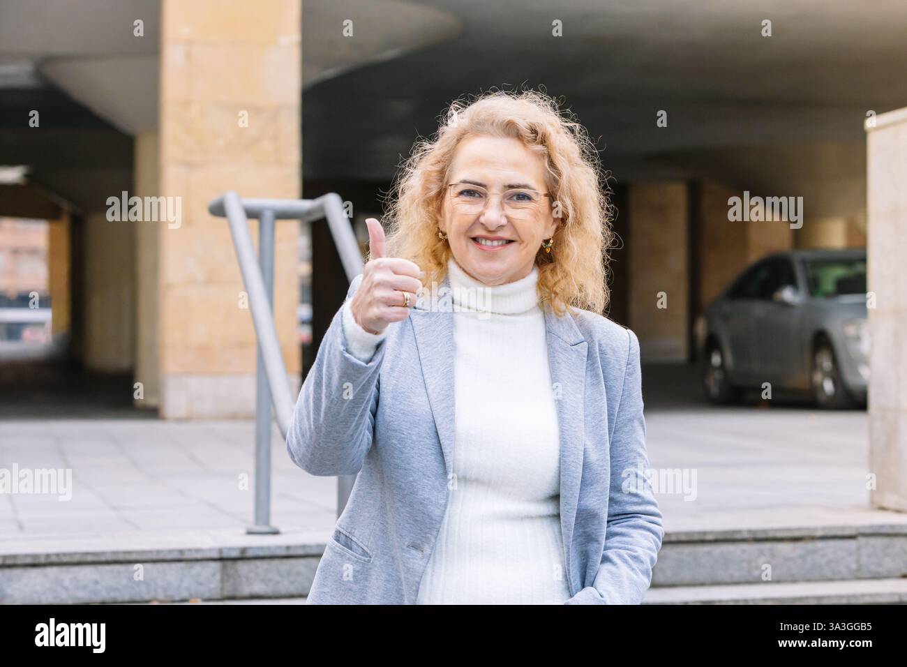 Cheerful mature businesswoman with curly blonde hair and glasses, wearing a light gray blazer and white turtleneck, smiling and giving a thumbs-up ges Stock Photo