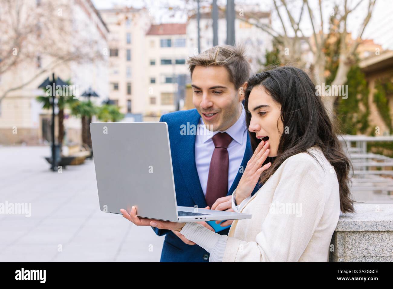 Two young business professionals in formal attire reacting with surprise while analyzing data on a laptop in an outdoor setting. concept of innovation Stock Photo