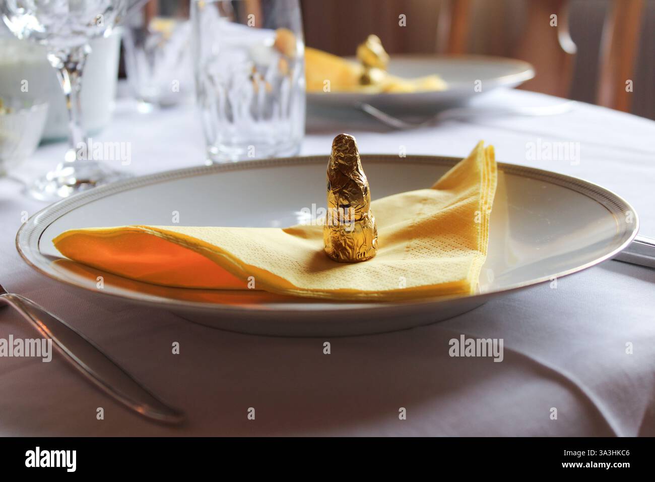 Elegant Easter table setting with a gold foil-wrapped chocolate bunny on a yellow napkin, placed on fine dinnerware. A festive and refined ambiance. Stock Photo