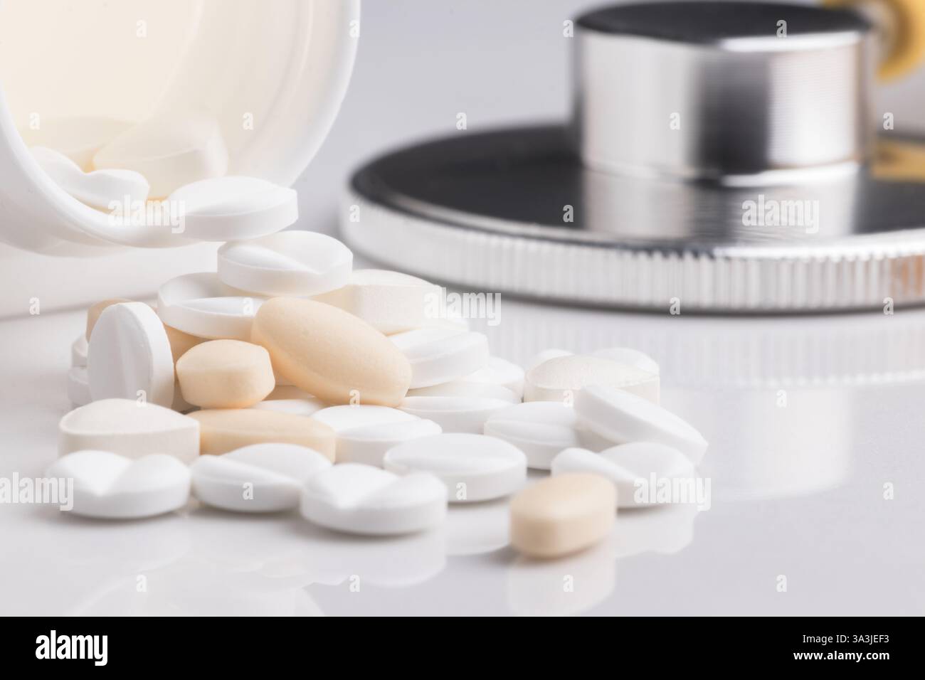 Close-up image of various white and beige medical pills scattered on a reflective surface, accompanied by a stethoscope. The image conveys themes of h Stock Photo