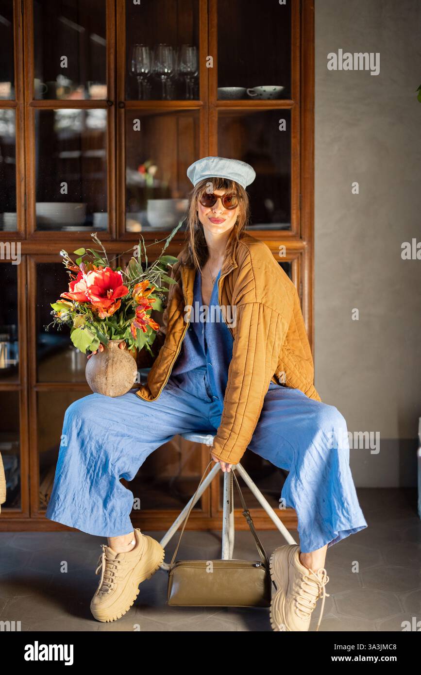 Trendy woman in a blue jumpsuit, brown quilted jacket, and beret poses confidently on a stool, holding a vase with vibrant flowers. The warm indoor setting enhances the chic and artistic mood Stock Photo