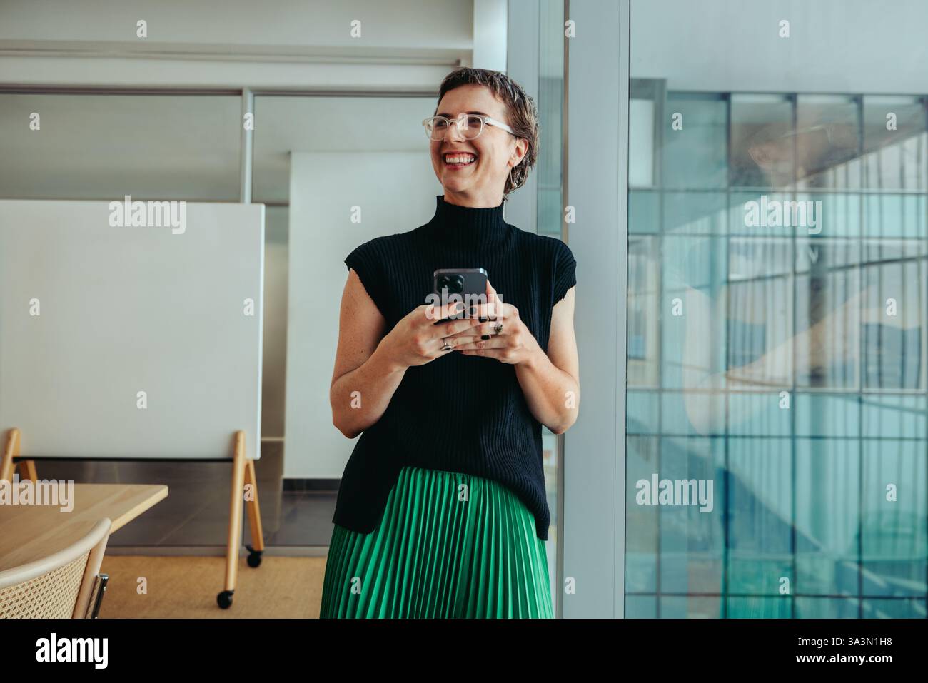 A non-binary entrepreneur smiling confidently while using her smartphone. She stands in a modern office environment, conveying themes of success, inno Stock Photo