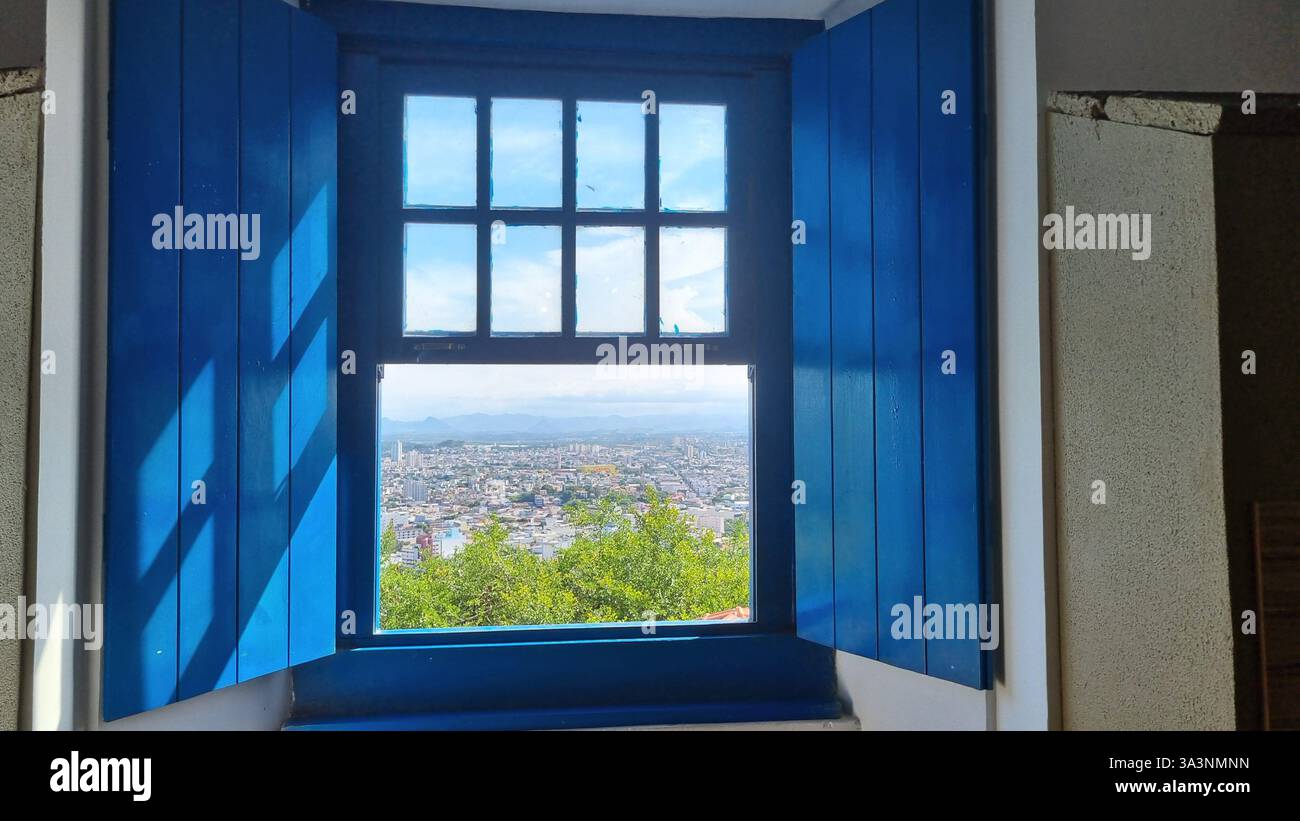 A beautiful open blue wooden window frames a breathtaking panoramic view of a cityscape below, with lush green foliage in the foreground and distant m - Smartphone Captured Stock Image