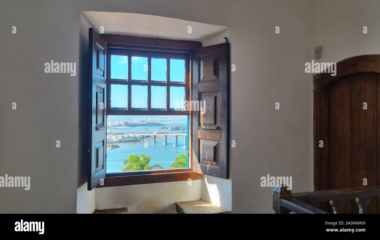 A beautiful open blue wooden window frames a breathtaking panoramic view of a cityscape below, with lush green foliage in the foreground and distant m - Smartphone Captured Stock Image