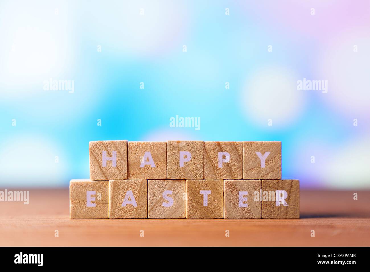Wooden blocks spelling HAPPY EASTER with colorful Easter eggs and flowers in the blurred background. A festive and cheerful scene symbolizing Easter Stock Photo