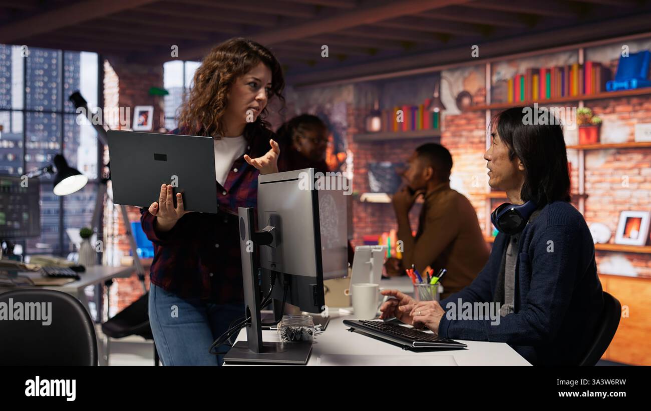 Startup intern holding laptop asks questions to senior technician coding advanced AI algorithm. Employee requests help from CTO manager in Silicon Valley workplace, camera B Stock Photo