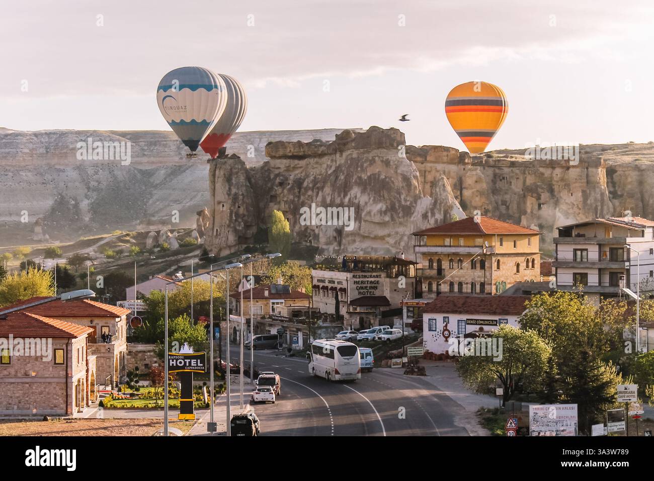 A group of colorful hot-air balloons floating close to the rocky landscapes of Goreme, Cappadocia, Turkey, at sunrise. A town in the foreground. Stock Photo