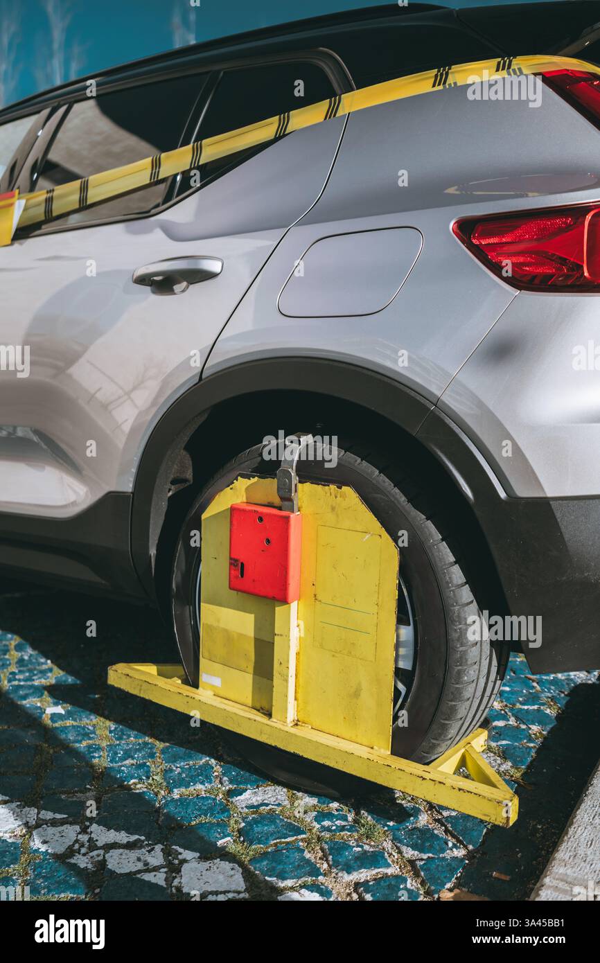 Close-up of a silver car immobilized by a yellow and red wheel clamp on cobblestone street, marked with caution tape, indicating parking violation or Stock Photo