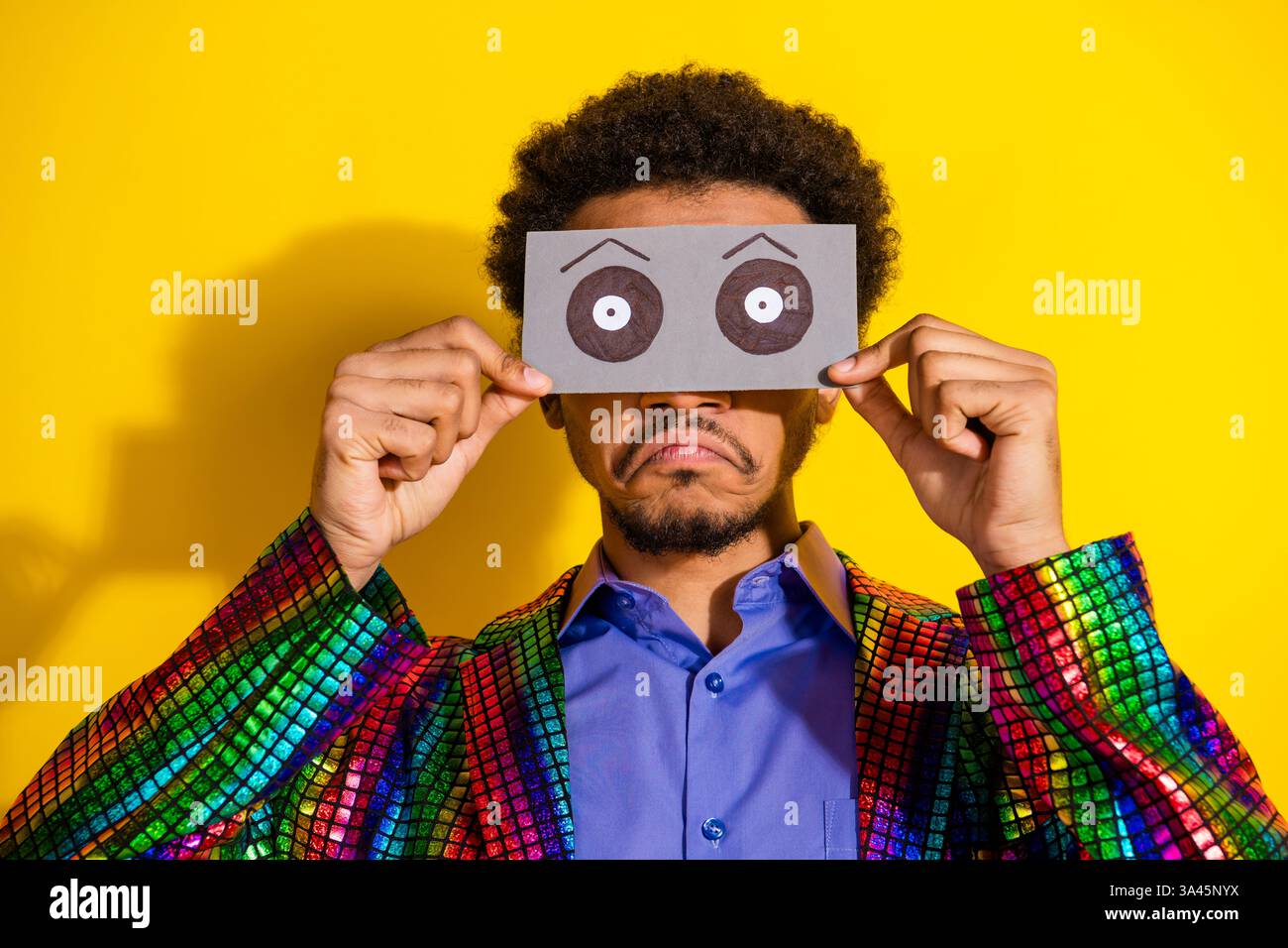 Young man in a colorful jacket holding a paper mask with drawn eyes, posing against a bright yellow background Stock Photo