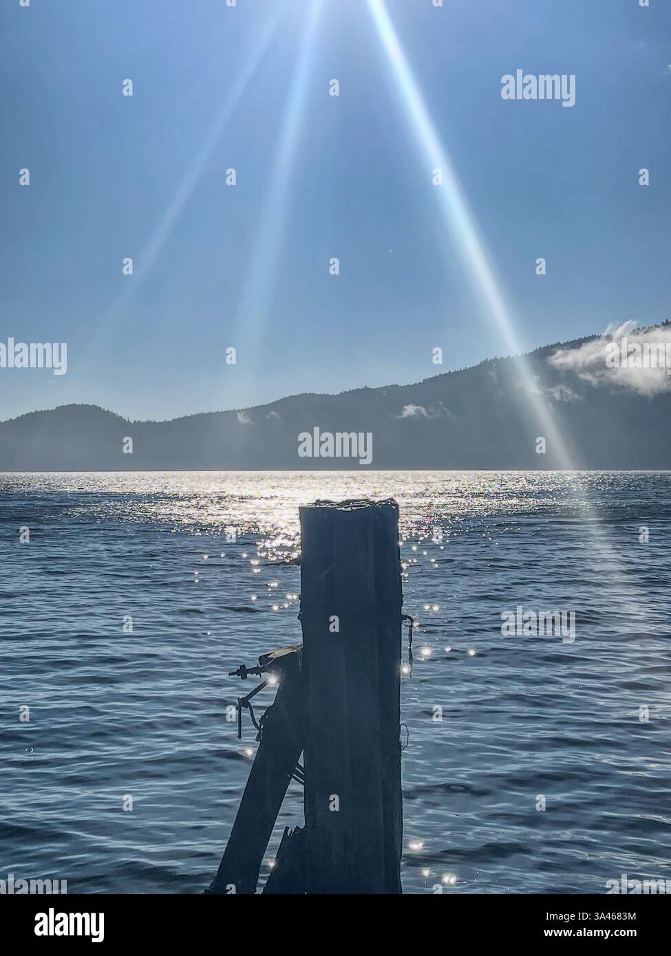 Sunlight streams down in beams over a calm body of water, creating sparkling reflections around an old wooden post, with distant mountains in the back - Smartphone Captured Stock Image