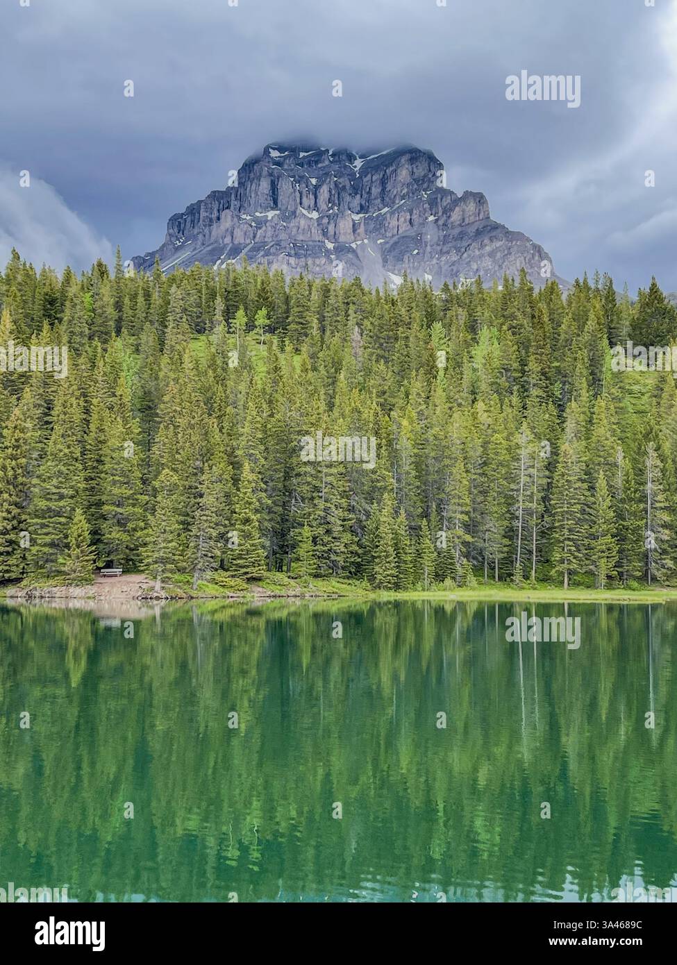 A striking mountain with rugged cliffs reflected in a green alpine lake, surrounded by a dense evergreen forest, under a cloudy sky in a remote wilder - Smartphone Captured Stock Image