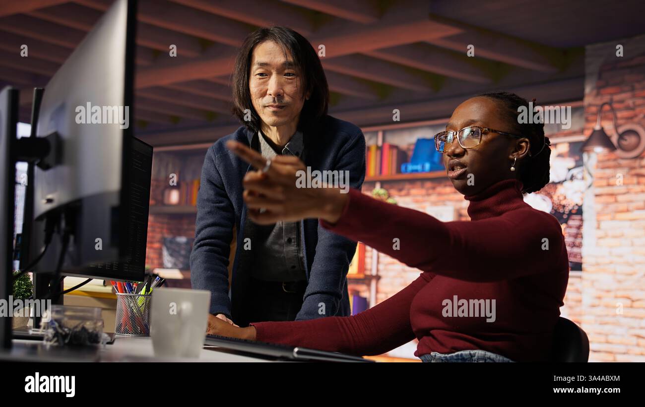 Man in AI startup office CTO position overseeing software engineer implementing machine learning. Artificial intelligence tech venture supervisor helps employee fix code bugs, camera B Stock Photo