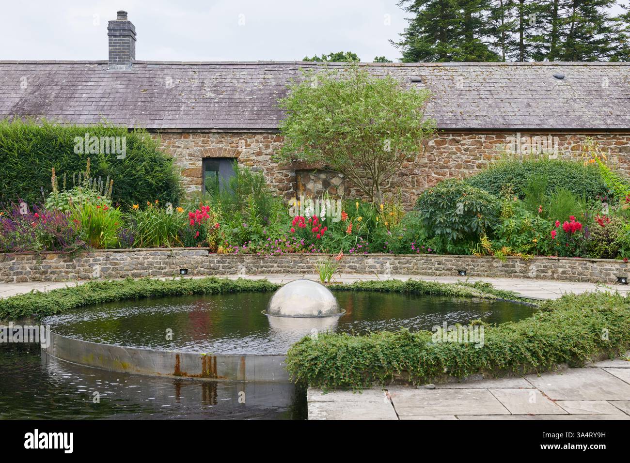 Large round water foreground feature with silver sphere in the centre, in a garden with a barn building in the background in South Wales, UK Stock Photo