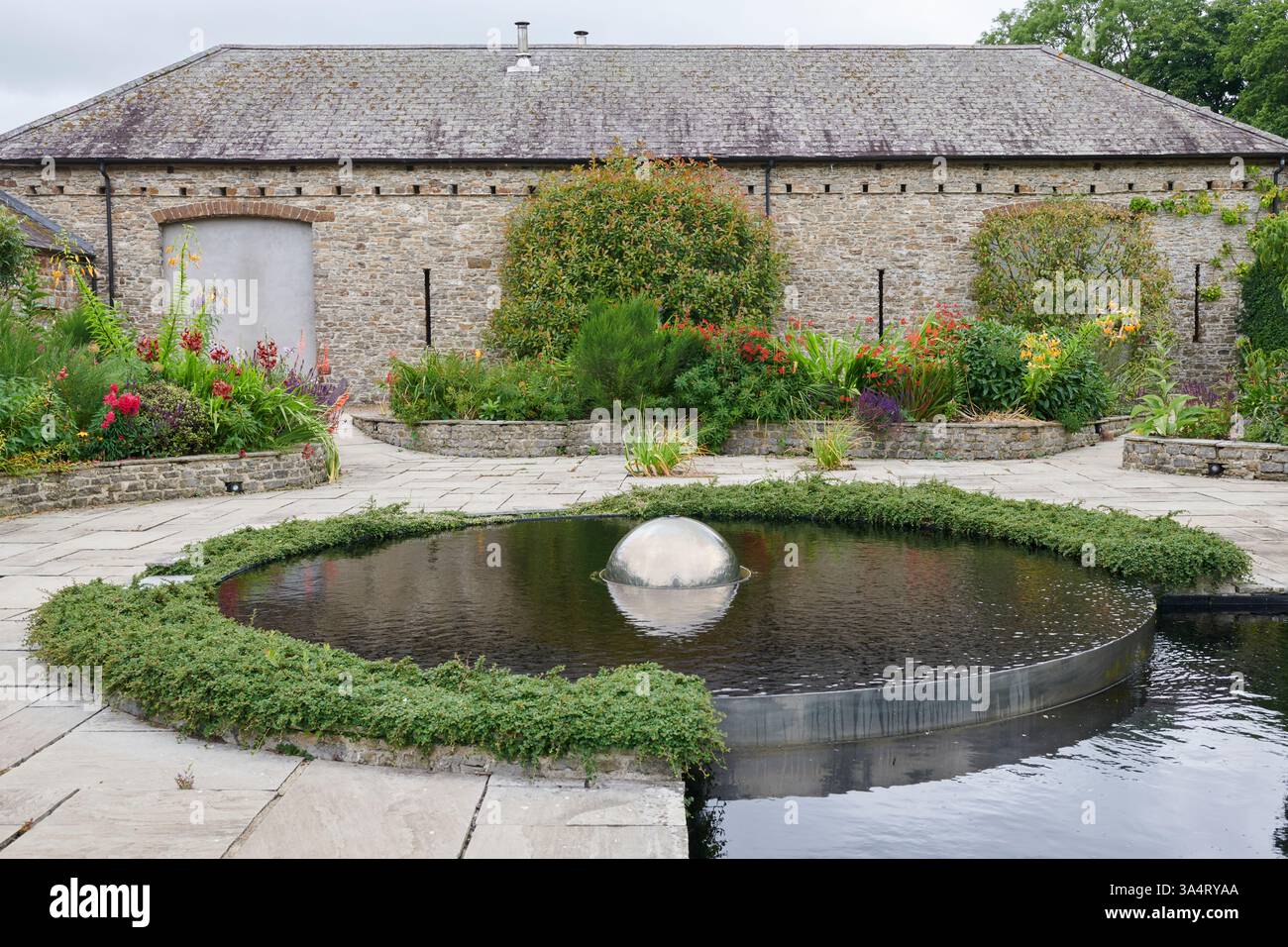 Large round water foreground feature with silver sphere in the centre, in a garden with a barn building in the background in South Wales, UK Stock Photo