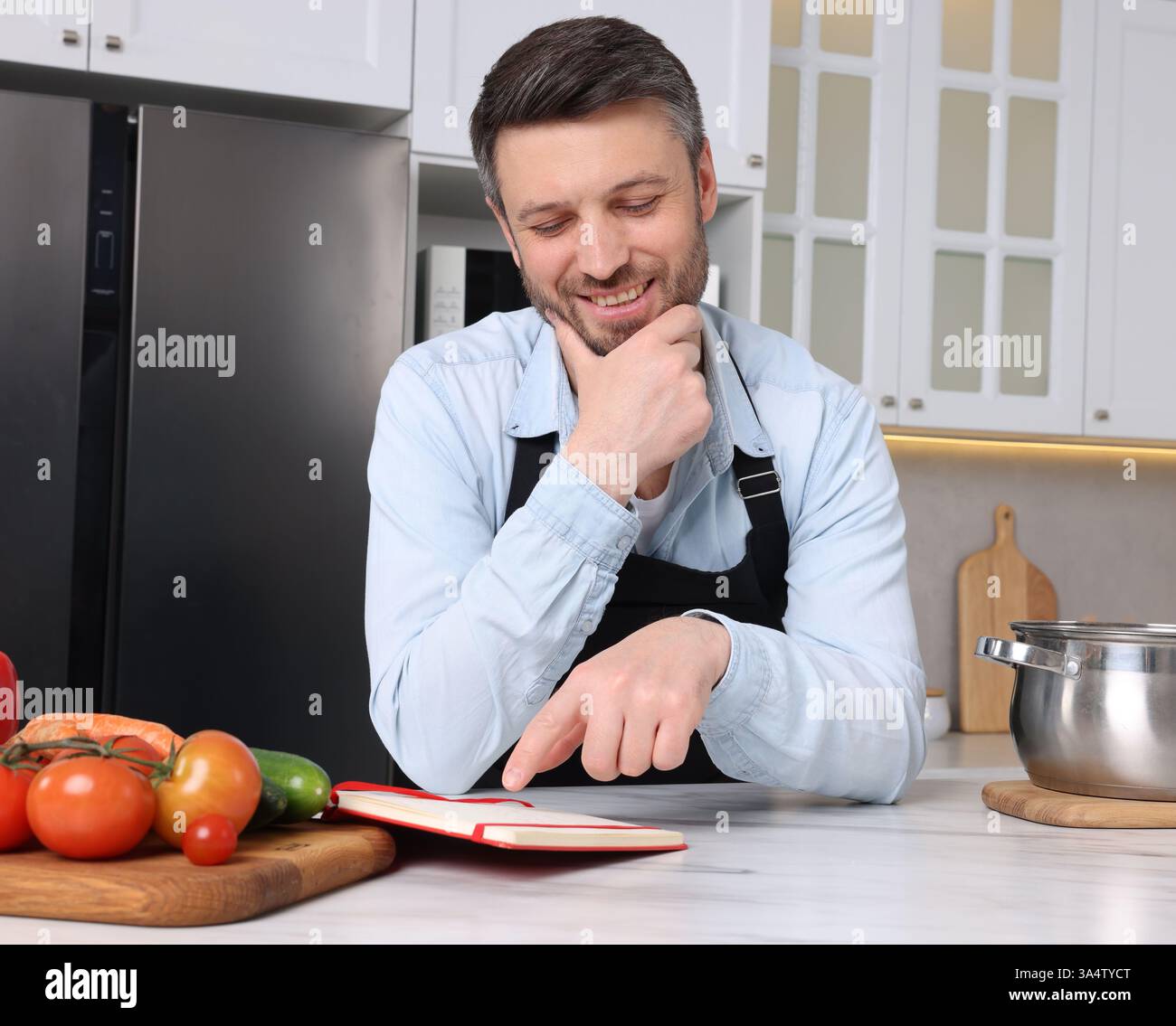 Adult man reading recipe book at white marble table in kitchen Stock Photo