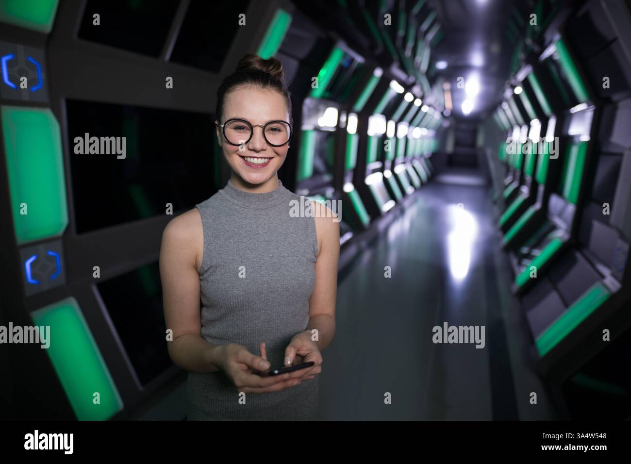 Smiling woman using smartphone in futuristic office corridor with green lights, copy space Stock Photo