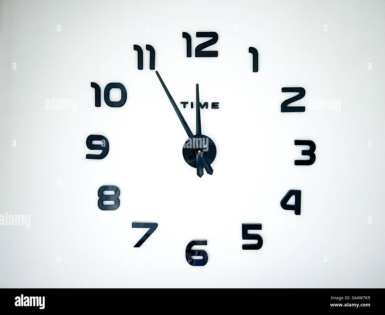 A minimalist wall clock with black hands and numbers on a white background, showing 5 minutes to 12 o’clock, symbolizing urgency, countdown. - Smartphone Captured Stock Image