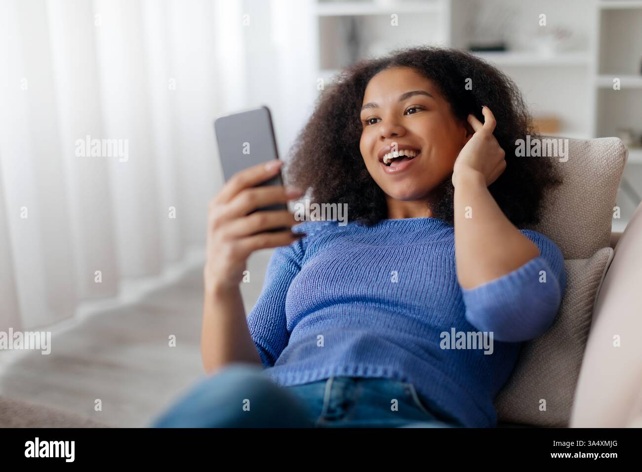 Young African American woman lying on couch, laughing while looking at smartphone Stock Photo
