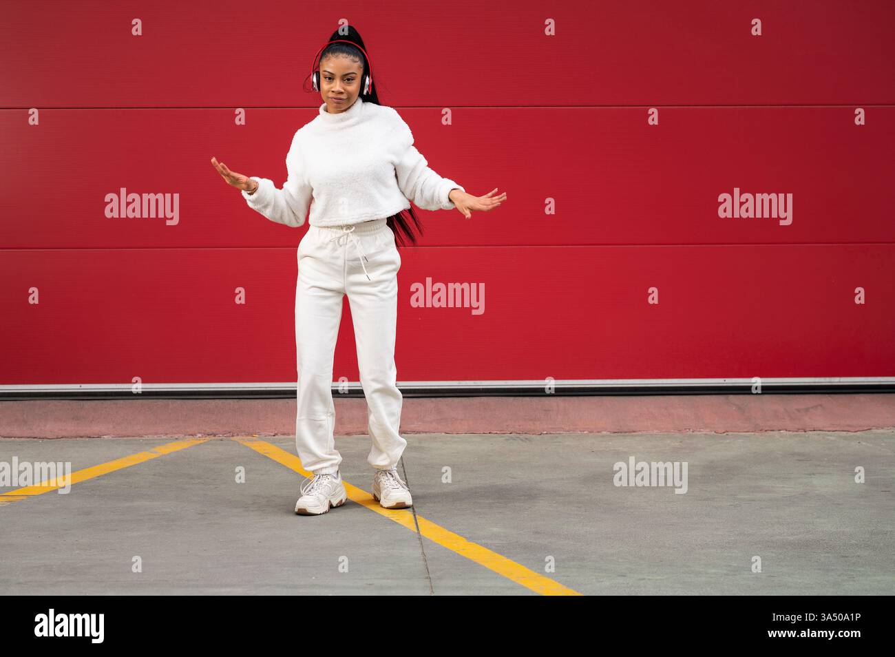 Energetic street dancer portrait of a smiling African American woman with long black hair wearing headphones, dancing in front of a vibrant red wall. This urban image is perfect for promoting hip hop, street style, and music lifestyle campaigns. Stock Photo