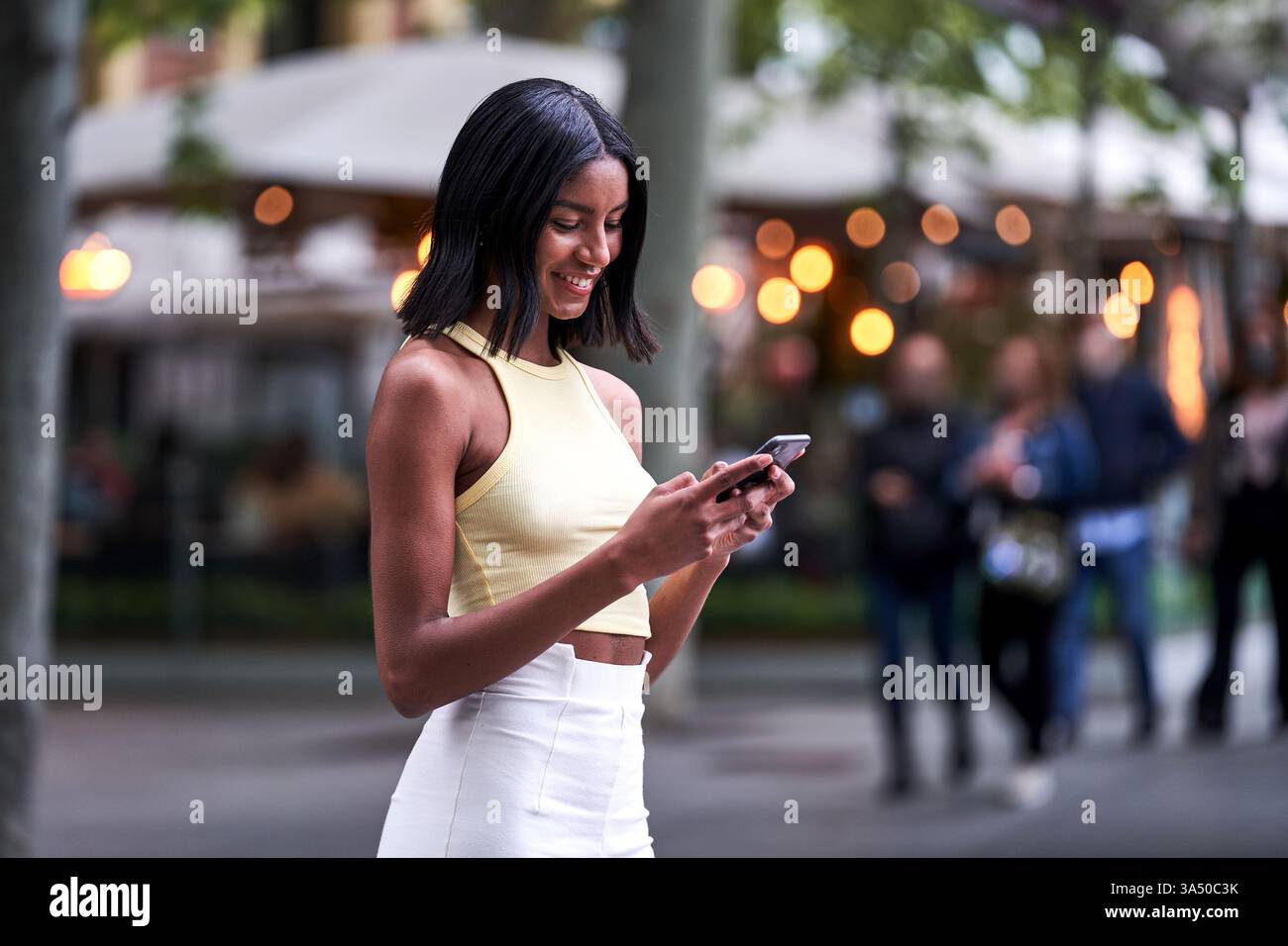 Young Black woman in stylish clothes texting on smartphone on a sunlit city street. The scene captures modern connectivity, urban lifestyle, and weekend leisure. Ideal for tech, social media, and fashion-related content. Stock Photo