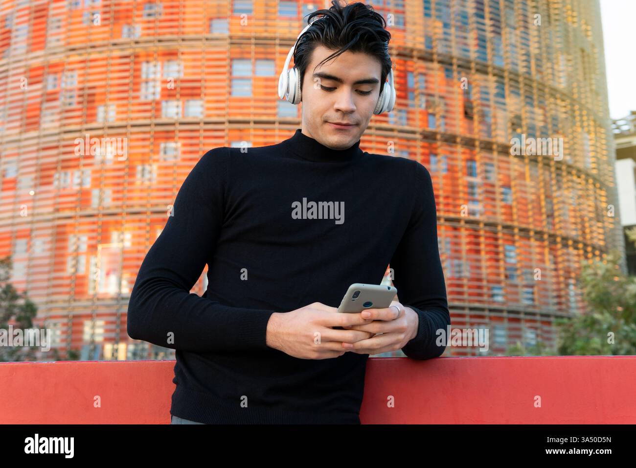 Hispanic man wearing headphones and using a smartphone on a city street. A modern urban lifestyle image suitable for music, mobile apps, and tech connectivity campaigns. Stock Photo