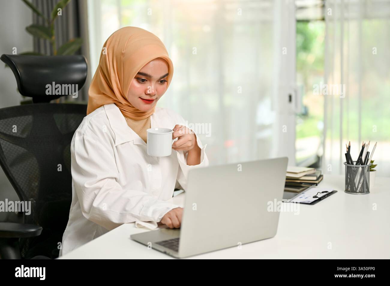 Asian Muslim woman in hijab sitting at a desk working on a laptop with ...