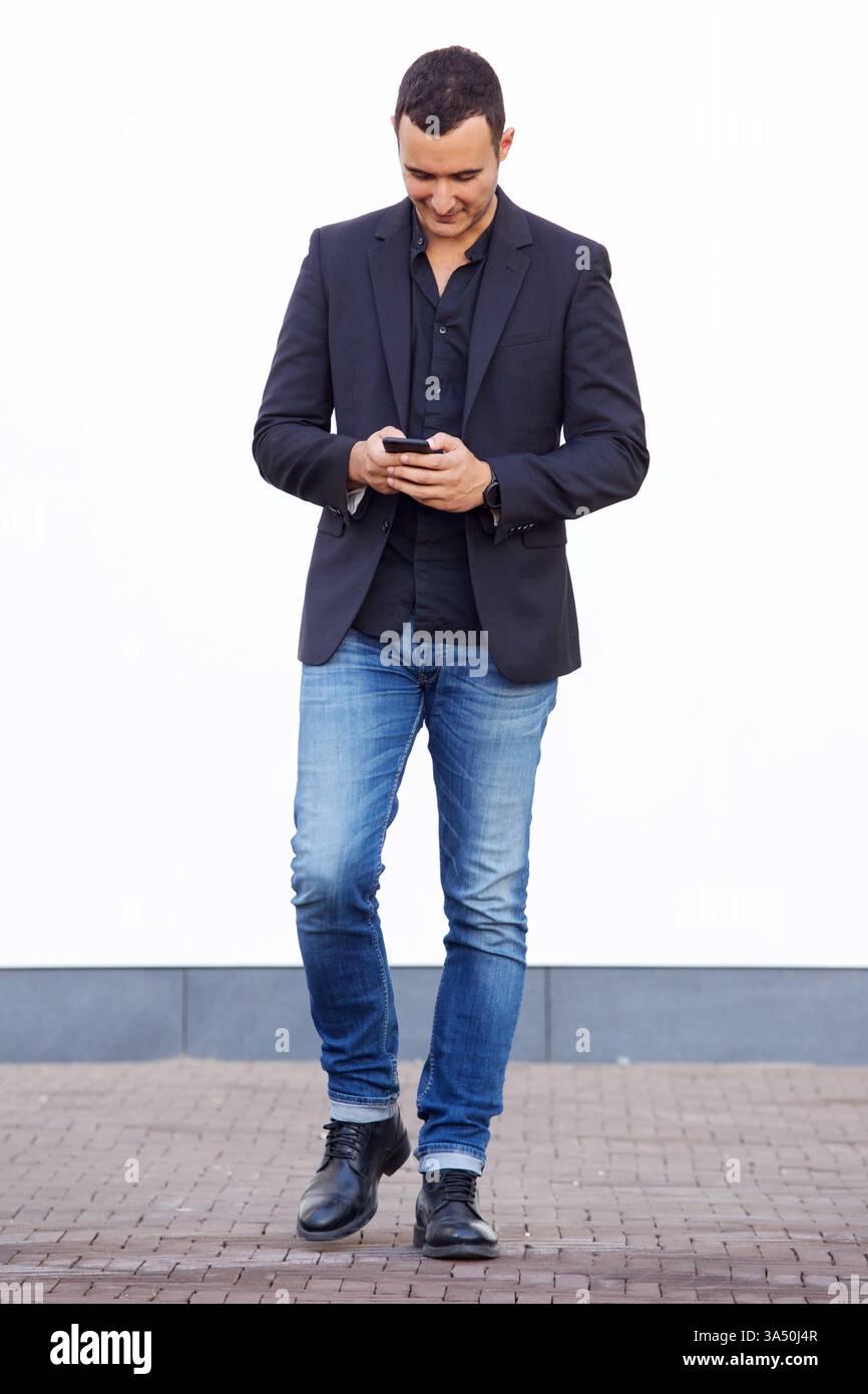 Hispanic businessman using smartphone walking against white wall on street during daytime Stock Photo