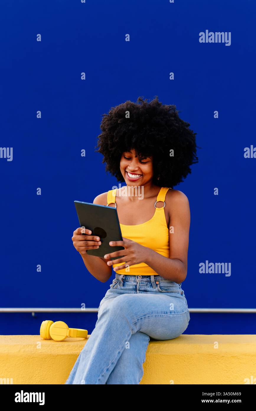 Smiling Black woman with an Afro using a tablet while sitting cross legged on a yellow fence beside a blue wall in an urban street setting. This lifestyle image reflects tech use and contemporary style. Stock Photo