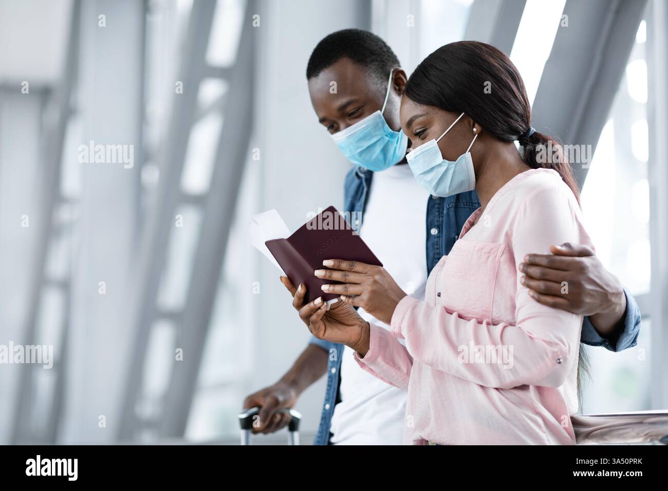 Checking Flight Information. Black Couple In Protective Masks Waiting For Departure In Airport, Holding Passports, Looking Boarding Time Stock Photo
