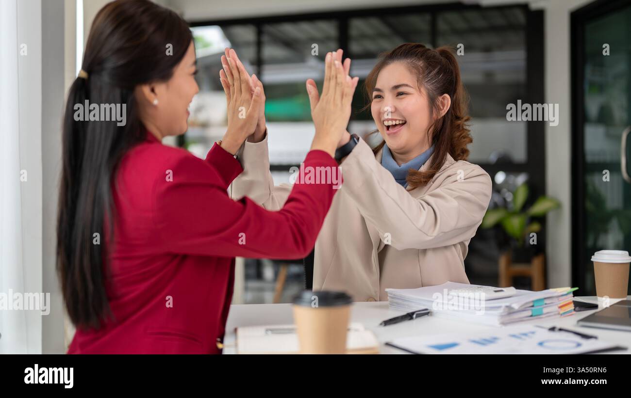 Two Asian businesswomen celebrate with a high five in a bright office ...