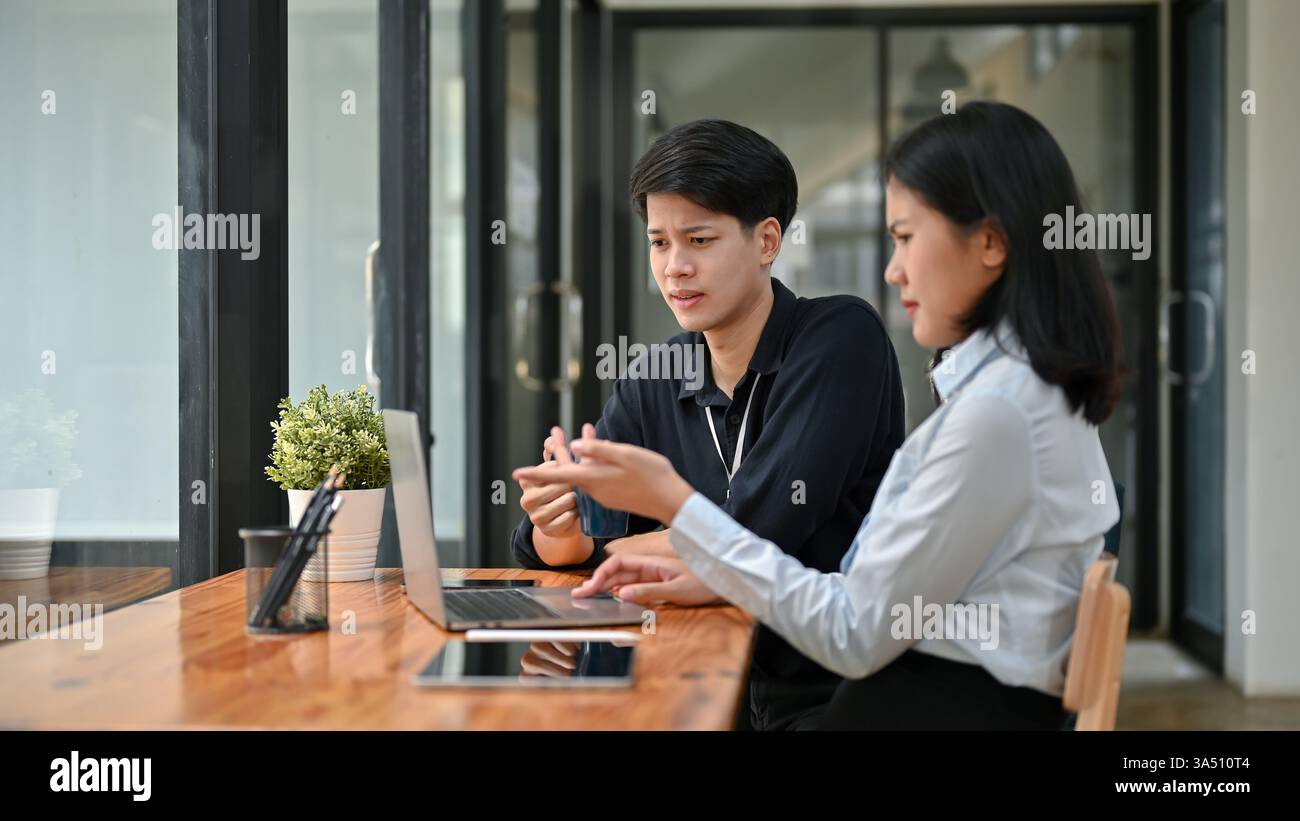 Confused Asian male and female office workers pointing at laptop screen having problem with their project while sitting together at table in cafe Stock Photo