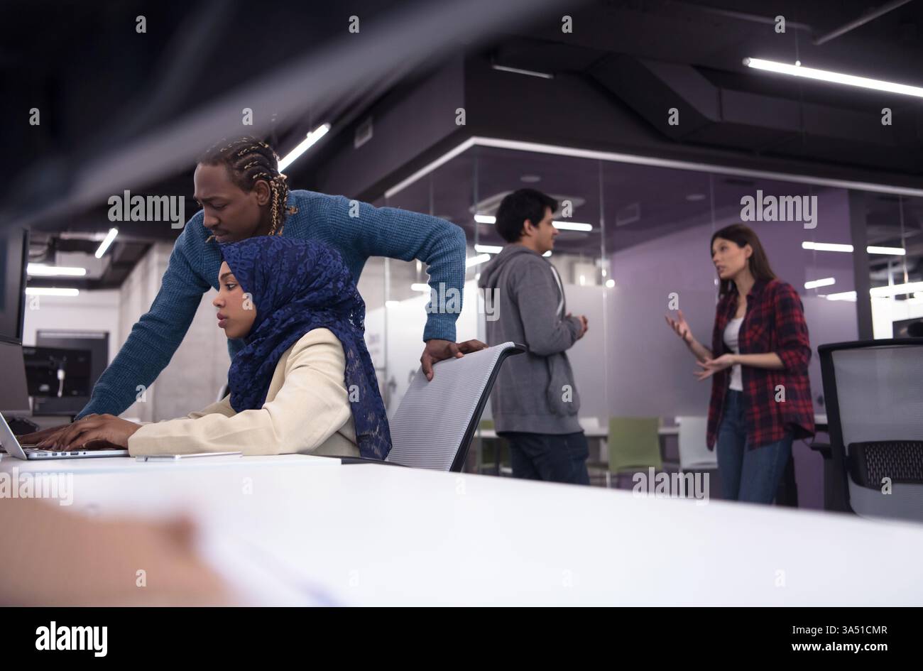 Black Muslim female software developer collaborates with a male colleague, coding on a laptop in a modern office. This diverse tech scene emphasizes teamwork and software development. Stock Photo