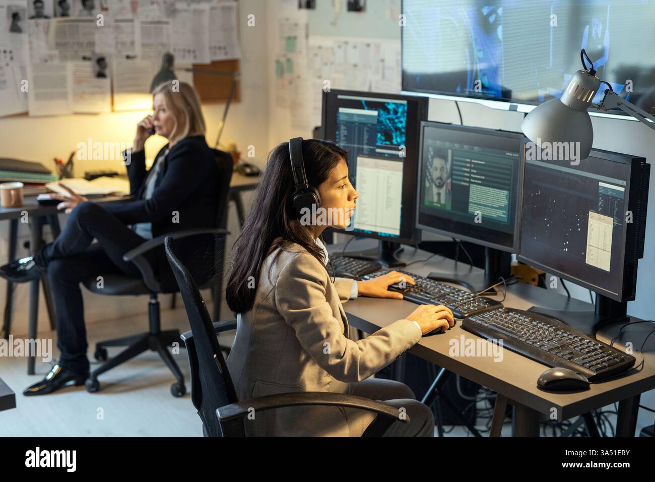 Serious female intelligence officer sits at a desk in a modern office, focused on computer monitors and data. This high tech scene suits stories about cyber security, crime investigation, and government agencies. A professional stock photo for law enforcement, intelligence, and data processing topics. Stock Photo