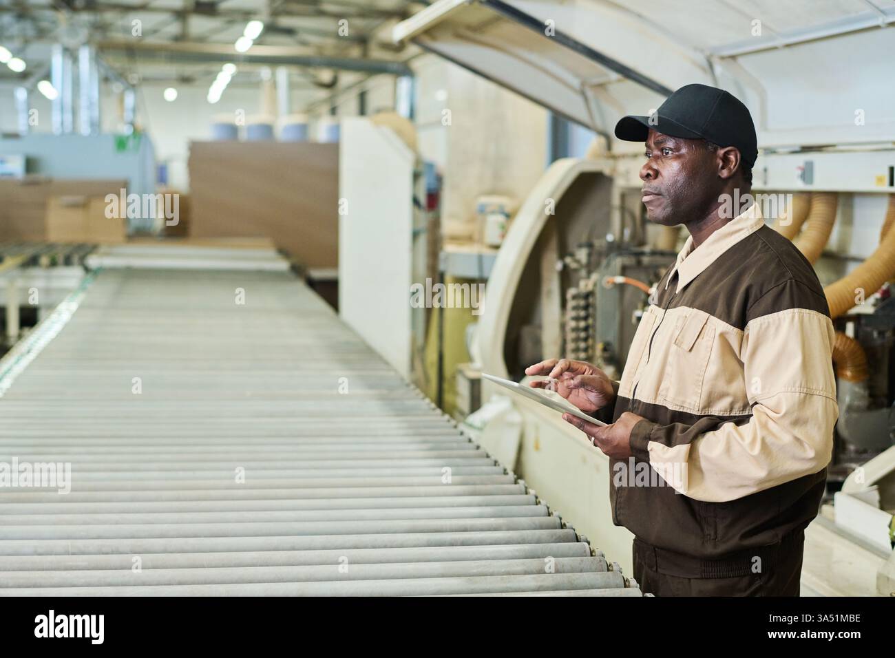African foreman in uniform monitors factory machinery using a digital tablet. This professional production scene highlights leadership and technical oversight in a modern manufacturing environment. Ideal for articles about manufacturing, engineering, and workplace efficiency. Stock Photo
