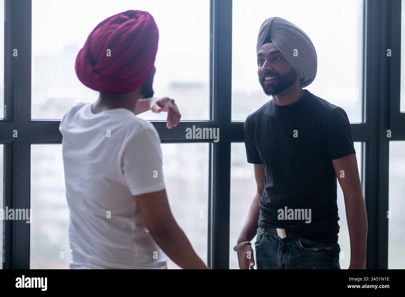 Two Indian men wearing turbans talking near a window in a home setting. The scene reflects everyday conversation and cultural diversity, suitable for lifestyle or community features. Stock Photo
