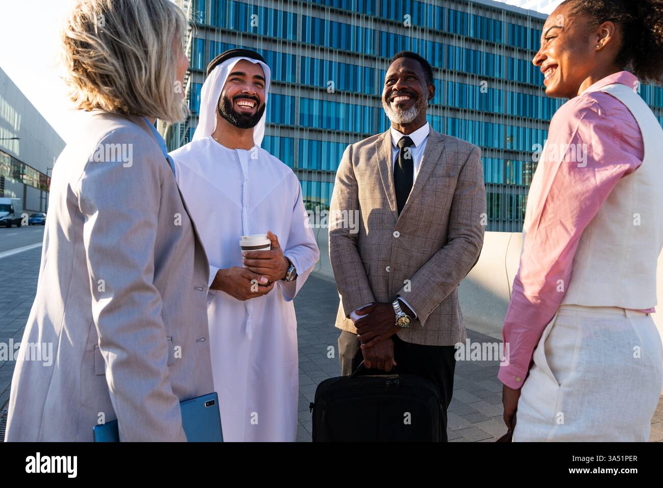 A diverse group of business professionals meets outdoors in an urban setting for a group discussion and team bonding. The scene highlights multicultural collaboration and networking against a modern city backdrop. Stock Photo