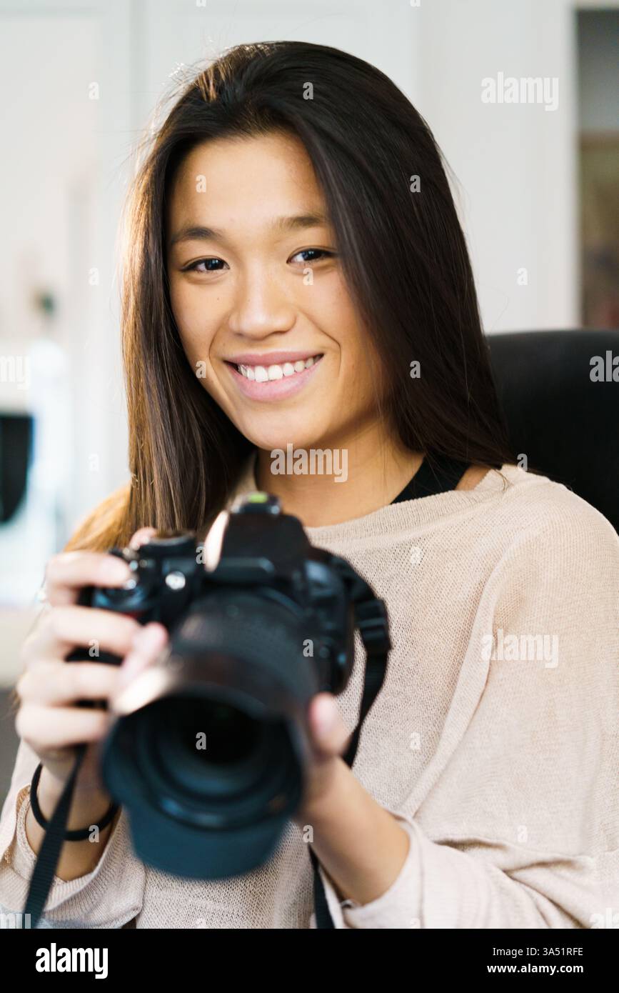 Smiling Asian female holding camera sitting on chair in workplace Stock Photo