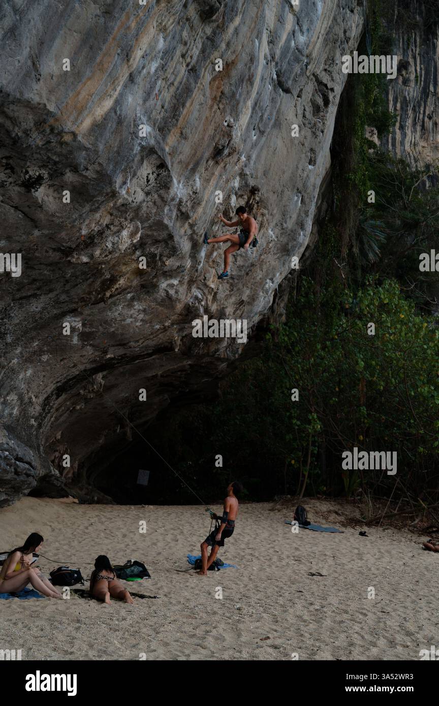 A climber scales a steep limestone cliff, a belayer manages the rope below, beachgoers relax on the sand Stock Photo