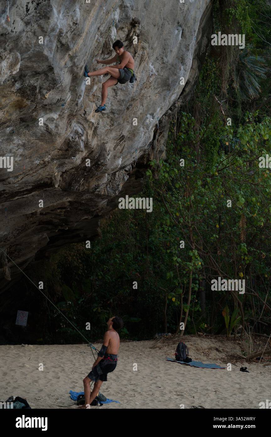 Young rock climber ascends a steep limestone cliff with a belayer ensuring safety from below Stock Photo