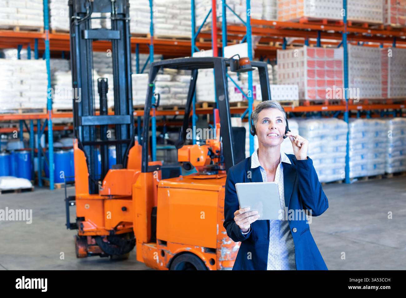 Coordinating logistics, warehouse manager using tablet and headset with forklift behind, copy space Stock Photo
