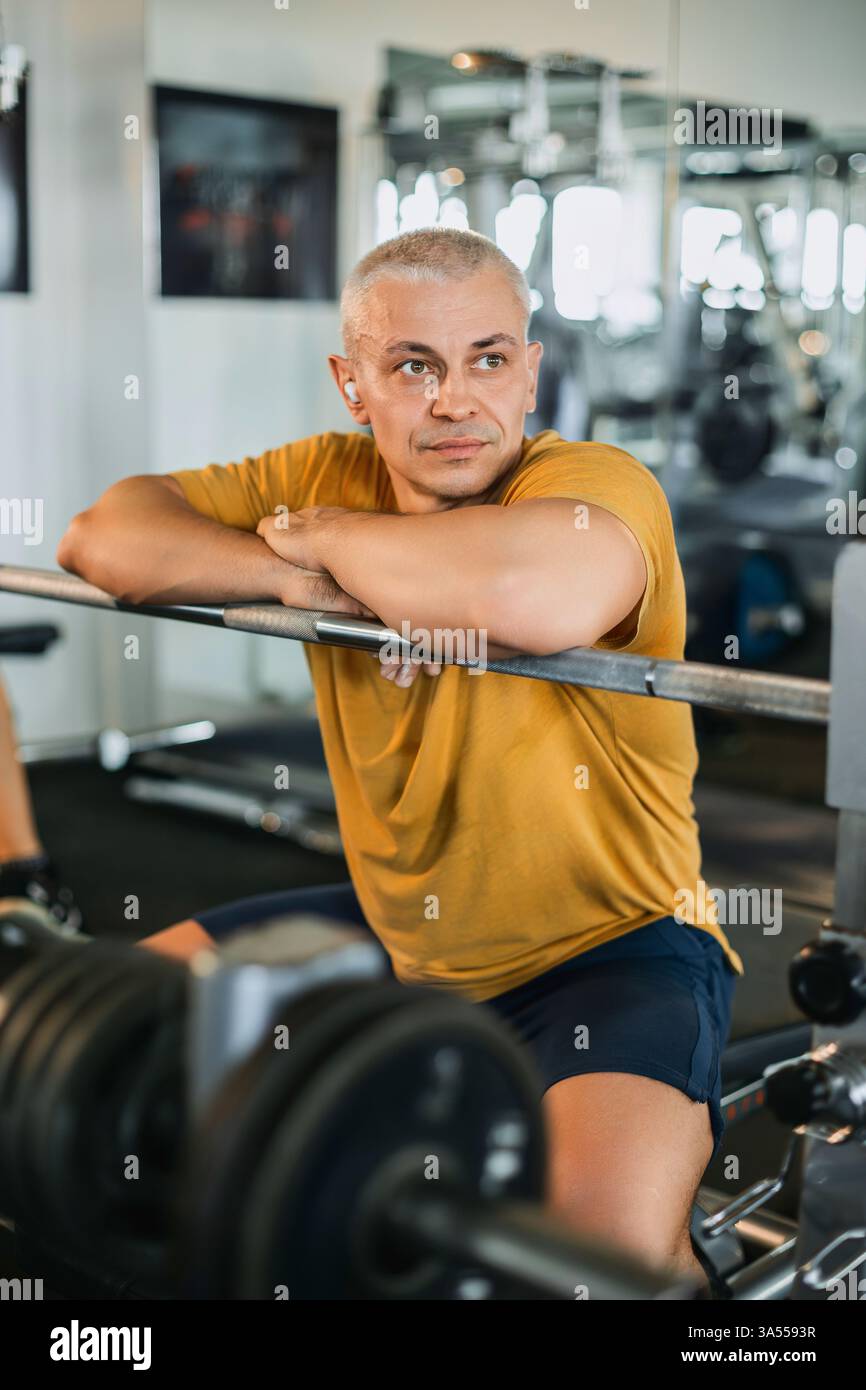 Fit mature man thoughtfully resting on barbell bench in gym setting Stock Photo