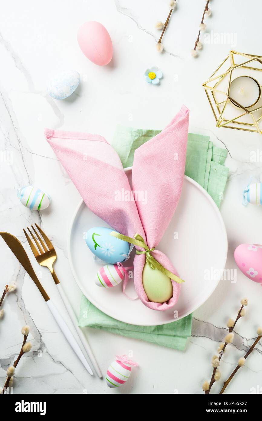 Easter table setting on white background with spring decorations. White plate, cutlery and colored eggs. Flat lay. Stock Photo