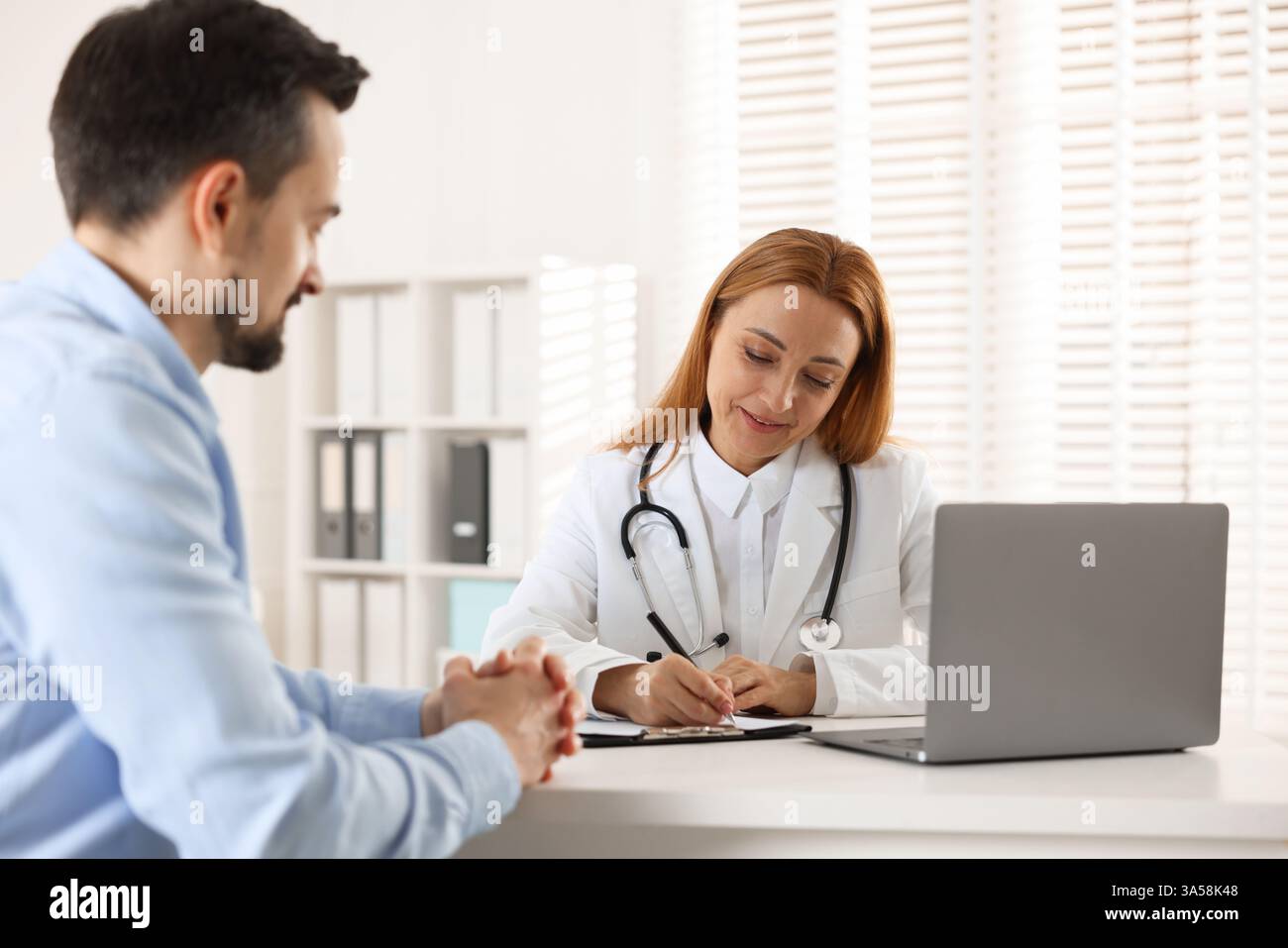 Cardiologist taking notes while consulting patient at table in clinic Stock Photo
