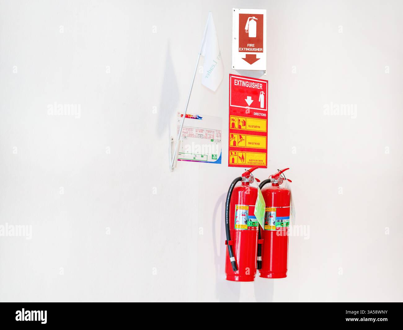 A red fire extinguisher and flags placed below an exit sign on a white wall Stock Photo