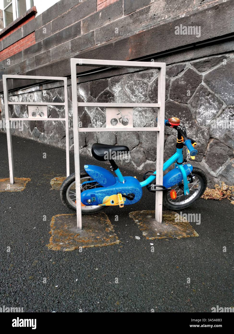 A vibrant blue child's bicycle is parked securely between two metal bike racks on a stone wall-lined street. - Smartphone Captured Stock Image